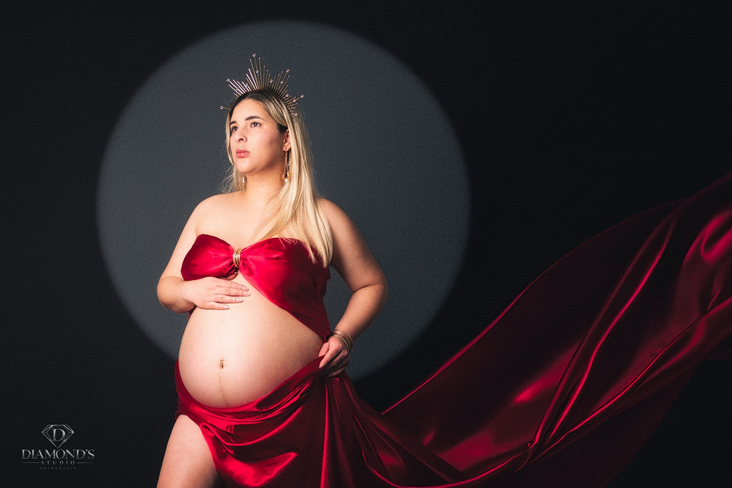 Sesión de maternidad: de pie con satén rojo y corona sobre fondo oscuro